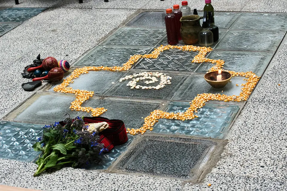 An altar laid out on the floor: a chakana, the Andean stepped cross, outlined in dried yellow corn around a spiral of white corn, with a lit candle in a clay bowl, maracas, bottles, a red woven sash and a bunch of fresh herbs with blue flowers