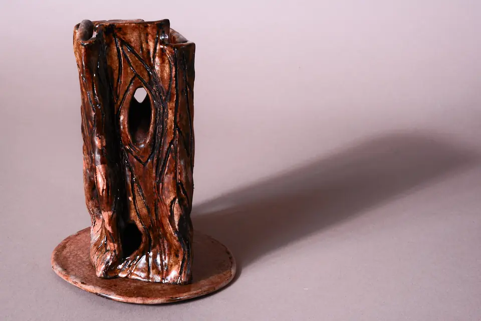 Tall brown-glazed vessel by gabmary with grooved lines and an almond-shaped opening through its upper half, standing on a round crackle-glazed disc; light from the left throws a long shadow across a pale lilac backdrop