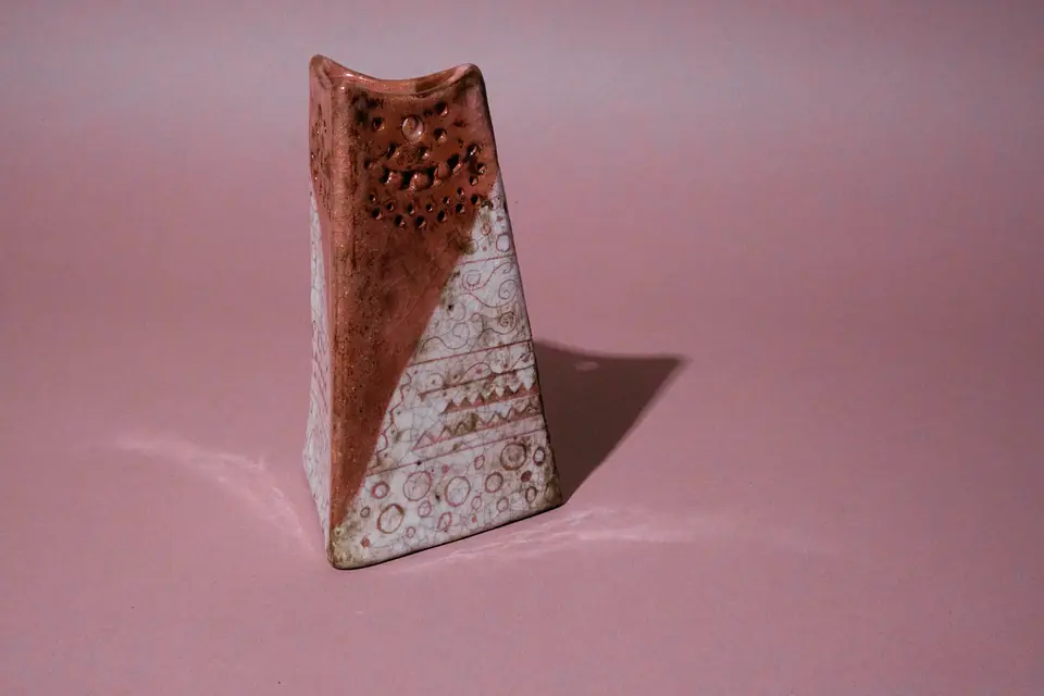 Tapered, angular vessel by gabmary with a curved rim: rust-pink glaze dotted with dimples and small holes near the top, crackled white below with pink spirals, zigzags and circles; hard shadow to the right with a bright spot inside it, on a pink backdrop