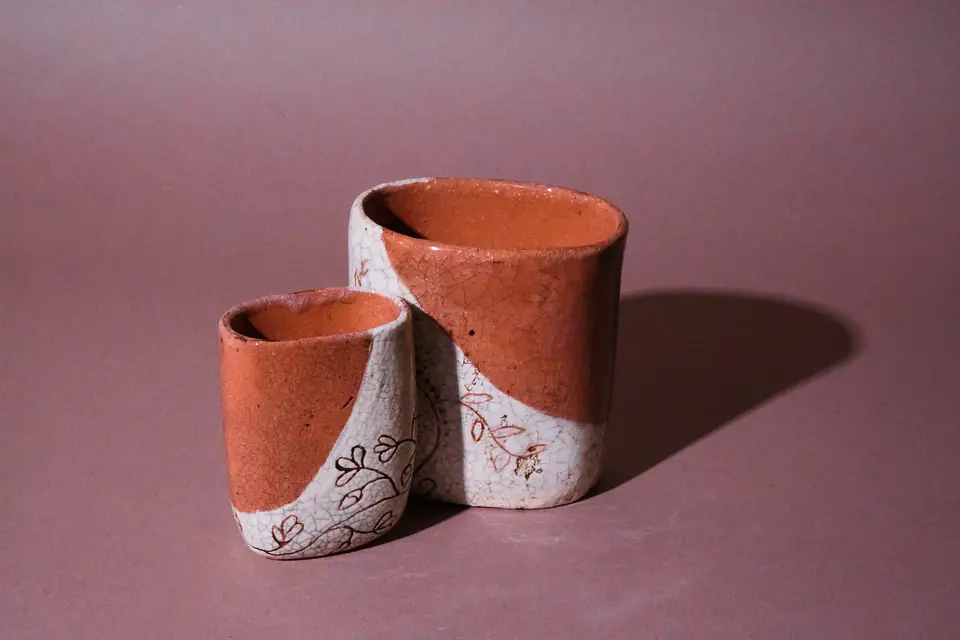 Two vessels by gabmary, a small one in front of a larger one, each glazed orange and crackled white along a curved line, with leaf-and-vine linework on the white; hard shadow to the right on a dusty mauve backdrop